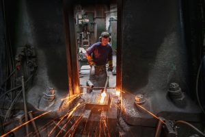 Apprentice engineer forging steel parts in hammer press in industrial forge